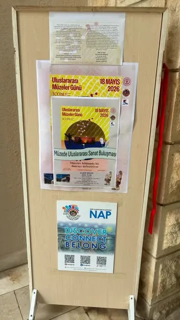 Ahşap bir panoya asılmış afişler: üstte “Uluslararası Müzeler Günü, 18 Mayıs 2026” ve “Müzede Uluslararası Sanat Buluşması” yazan afiş, altında Yeni Alanyalılar (New Alanyans Platform, NAP) platformunun afişi ve karekodlar.