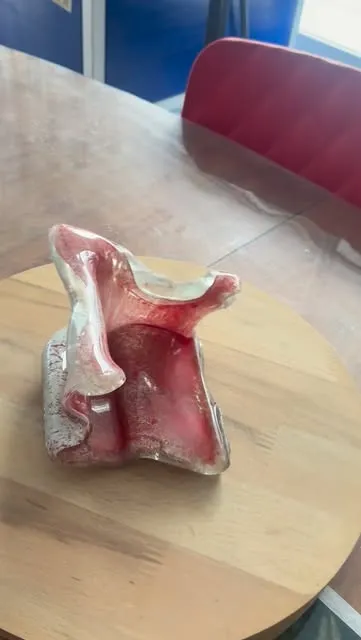 Yuvarlak, açık renk bir ahşap tabla üzerinde duran, kenarları kıvrılmış pembe-kırmızı serbest biçimli cam çalışma.