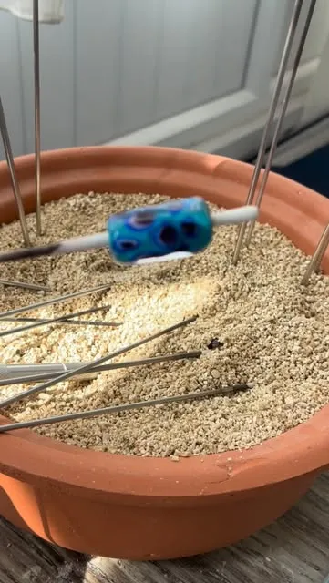 Pişmiş toprak bir kapta, vermikülitin üzerinde çelik çubuğunun ucunda soğumaya bırakılmış mavi-turkuaz lampwork cam boncuk; yüzeyinde koyu mavi göz benzeri lekeler var.